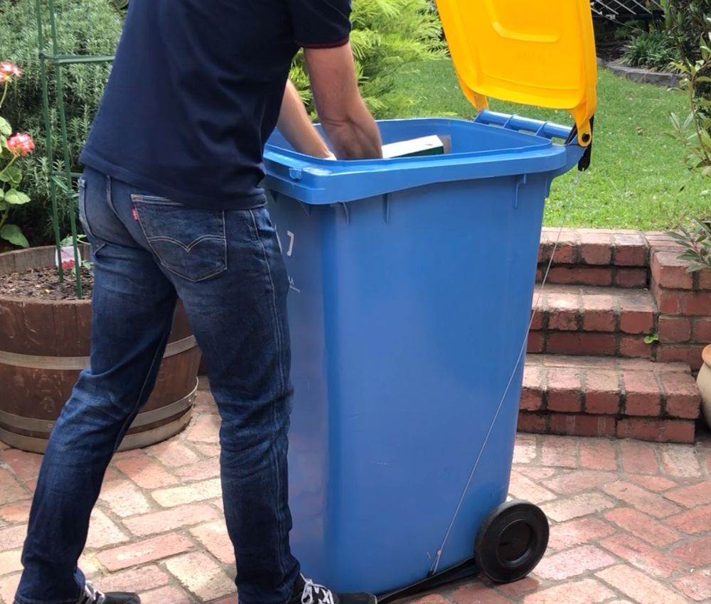 Person placing rubbish into a blue wheelie bin with a yellow lid, using the foot pedal of the bin lid opener