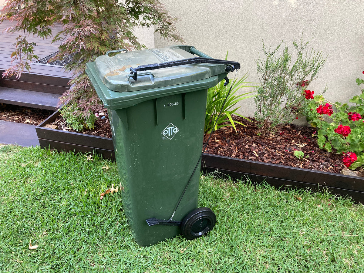 BLOP foot-operated bin lid opener installed on a green wheelie bin with a closed lid, showing the pedal, cable and lifting arm